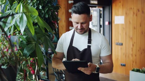 Hombre-guapo-en-delantal-disfrutando-de-flores-en-la-floristería-usando-tableta-sin-trabajo