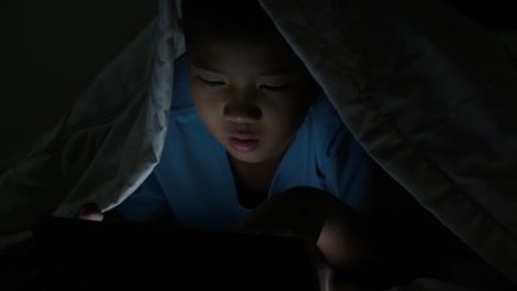 Little-boy-using-tablet-under-blanket-at-night-in-bed.