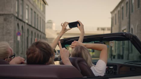 Zwei-schöne-Mädchen-und-zwei-stilvolle-Jungs-Chillen-und-Spaß-im-Cabrio-Auto,-mit-Smartphones,-Selfies,-Schießen-Video-und-Posting-auf-Social-Media.-Junge-Menschen-Reisen,-Sprechen