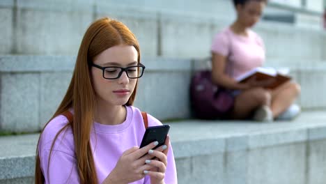 Sad-caucasian-female-teen-in-eyeglasses-messaging-social-networks-on-cellphone