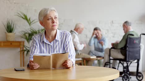 Rack-focus-of-three-elderly-people,-two-men-including-disabled-one-and-woman,-playing-cards-in-nursing-home.-Old-woman-with-short-grey-hair-reading-book-and-thinking-sadly-sitting-at-table-alone