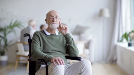 Tracking-medium-shot-of-disabled-elderly-man-in-eyeglasses-sitting-in-wheelchair-and-thinking,-then-turning-head-and-looking-sadly-at-camera-in-nursing-home