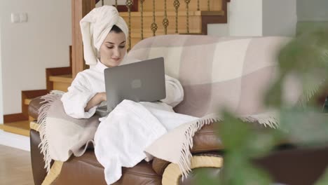 Beautiful-young-Caucasian-lady-in-bathrobe-and-hair-towel-lying-on-the-couch-and-typing-on-laptop.-Pretty-girl-using-social-media-at-home.-Cinema-4k-footage-ProRes-HQ.