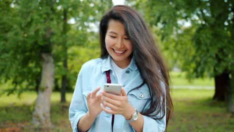 Atractiva-mujer-asiática-usando-teléfono-inteligente-en-el-parque-de-la-ciudad-tocando-la-pantalla-sonriendo