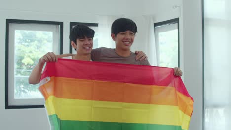 Porträt-Junge-asiatische-Homosexuell-Paar-Gefühl-glücklich-zeigt-Regenbogen-Flagge-zu-Hause.-Asia-LGBTQ+-Männer-entspannen-zu-schüchtern-Lächeln-blickend-in-die-Kamera,-während-Umarmung-im-modernen-Wohnzimmer-im-Haus-im-MorgenKonzept.