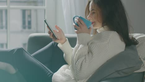 Beautiful-Young-Woman-Using-Smartphone,-Drinks-Tea-while-Sitting-on-the-Chair.-Sensual-Girl-Wearing-Sweater-Surfs-Internet,-Posts-On-Social-Media,-Shares-Pictures-while-Relaxing-in-Cozy-Apartment