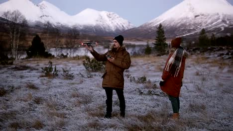 Una-pareja-caucásica-lanza-drone-en-el-campo-frente-a-las-montañas-con-picos-nevados-en-el-fondo.-Hombre-operando-el-helicóptero,-mujer-de-pie-cerca.-Campo-nevado-alrededor