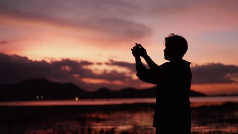 Silueta-de-retrato-asiático-hombre-usando-teléfono-inteligente-tomando-una-foto-en-la-playa-del-mar-hermosa-puesta-de-sol-de-verano,-luz-de-la-hora-dorada.