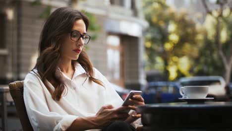 Geschäftsfrau-in-Brille,-weißes-Hemd.-Sie-sitzt-am-Tisch-mit-einer-Tasse-Kaffee-im-Straßencafé.-Durchsuchen-von-Nachrichten-auf-dem-Mobiltelefon.-Nahaufnahme