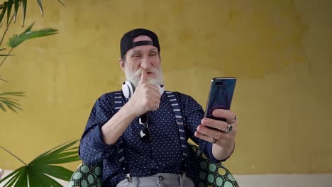 Front-view-of-funny-happy-attractive-bearded-senior-man-which-having-video-chat-on-phone-,sitting-in-chair-on-yellow-background