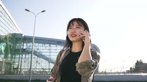 Hermoso-retrato-de-atractiva-joven-morena-asiática-con-el-pelo-largo-que-habla-por-teléfono-cerca-del-edificio-del-aeropuerto