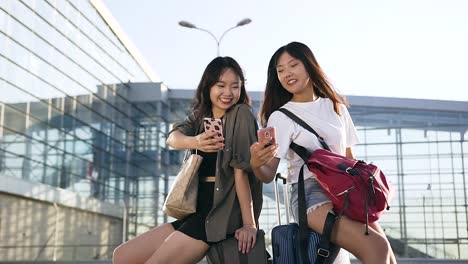 Front-view-of-two-charming-exuberant-asian-women-which-sitting-on-their-suitcases-and-using-smartphones-near-the-big-airport-terminal