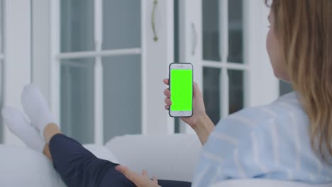 Over-shoulder-shot-of-woman-holding-a-mobile-phone-or-smartphone-with-a-vertical-green-screen-in-her-hand-at-home-lying-on-the-sofa.-Girl-tap-on-display-and-watching-content.