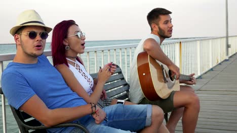 Tres-amigos-sentados-en-un-banco-con-el-mar-como-fondo-y-cantando-con-una-guitarra