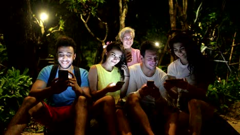 Grupo-de-personas-con-celular-inteligente-sentado-sobre-un-banco-en-el-parque-de-noche,-jóvenes-amigos,-hablando-de-redes-en-línea