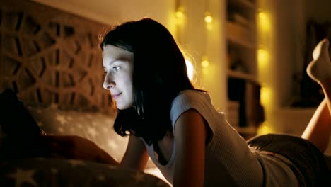 Young-smiling-woman-using-tablet-computer-for-sharing-social-media-lying-in-bed-at-home-at-night