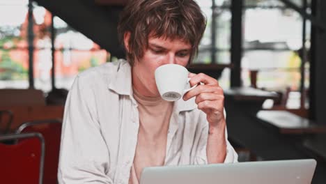 Young-attractive-cheerful-male-in-light-clothes-using-modern-portable-laptop-computer-and-drink-coffee-in-cafe.-Smiling-man-working-from-cafe-via-laptop,-social-networking-concept.-People,-communication-and-leisure.