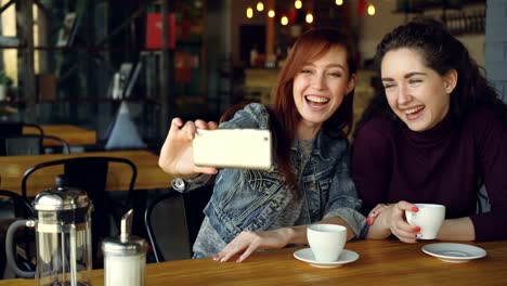 Hübsche-Mädchen-am-besten-Freunde-nehmen-Selfie-mit-Smartphone-dann-beobachten-Fotos-beim-Kaffeetrinken-im-Kaffeehaus.-Freundschaft,-social-Media-und-Spaß-Konzept.