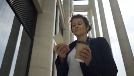 Low-angle-panning-shot-of-middle-aged-businesswoman-with-takeaway-coffee-cup-in-her-hand-text-messaging-on-smartphone-outdoors