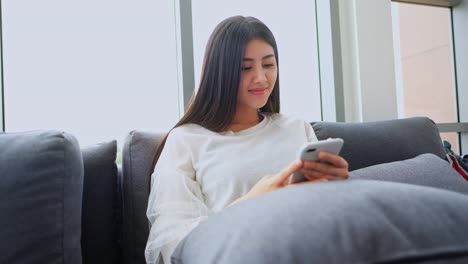 Asiatische-Frauen-im-Wohnzimmer-überprüfen-Smartphone-im-sozialen-Netzwerk.