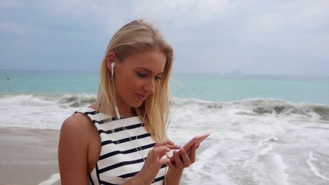 Young-beautiful-slim-woman-with-long-blonde-hair-in-black-and-white-dress-standing-on-the-coast-and-using-smartphone-over-background-at-storm-on-the-sea..-Girl-on-the-beach-touching-screen-and-smile.