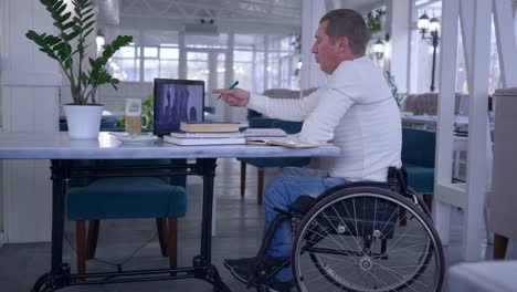 online-training,-student-disabled-in-wheelchair-making-notes-in-notebook-during-distant-education-sitting-at-table-with-laptop-computer-in-cafe