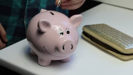 On-the-table-there-is-a-piggy-bank-in-the-form-of-a-pink-pig.-Lying-wallet-and-scattered-coins.-A-man-collects-coins-from-the-table,-puts-them-in-a-piggy-bank-and-closes-the-wallet.