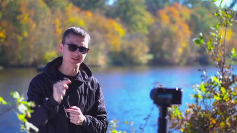 Blogging-young-man-in-sunglasses-stand-next-to-camera-at-nature-showing-thumbs-up-and-saying-goodbye-for-subscribers-slow-motion