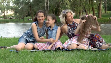 Happy-same-sex-family:-two-mothers-and-two-little-daughters-play-and-have-fun-sitting-on-the-grass-in-the-Park-at-sunset-on-a-summer-day-against-the-river.-Family-vacation-in-nature.-4K.