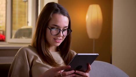 Retrato-en-primer-plano-de-joven-atractiva-mujer-caucásica-en-gafas-usando-la-tableta-sentada-en-el-sofá-en-el-interior-del-apartamento