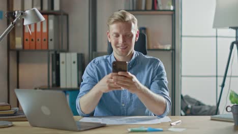 Creative-Entrepreneur-Sitting-at-His-Desk-Works-on-a-Laptop-in-the-Stylish-Office,-Uses-Smartphone-for-Social-Media-Apps,-Emailing-Business-Associates,-Reading-News,-Browsing-through-Internet