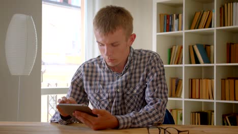 Portrait-of-caucasian-businessman-working-with-modern-tablet-attentively-on-bookshelves-background.