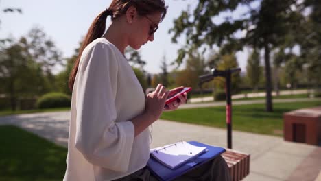 Stylish-caucasian-woman-in-a-white-blouse-and-glasses-is-typing-a-message-on-the-phone.-Electric-scooter,-business,-online-purchase