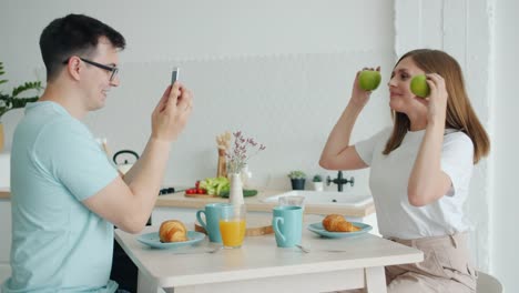 Chica-posando-con-fruta-mientras-que-el-chico-tomando-fotos-con-el-teléfono-inteligente-en-la-mesa-de-la-cocina