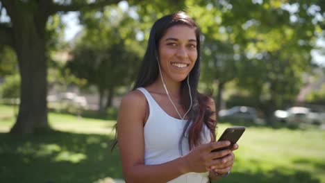 Cheerful-young-brunette-mixed-race-woman-with-earphones-in-her-ears-look-at-camera-and-smiles-white-texting-messages-on-mobile-phone-in-the-park