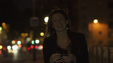 Caucasian-modern-woman-wearing-flower-dress,-black-jacket-and-red-hair-walking-through-the-street-and-writing-a-text-message-on-smartphone-and-laughing-at-camera-by-night.-Paris-4K-UHD.-Slow-Motion.