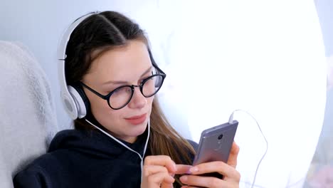 Woman-listens-music-in-headphones-on-smartphone-sitting-in-armchair-at-home.