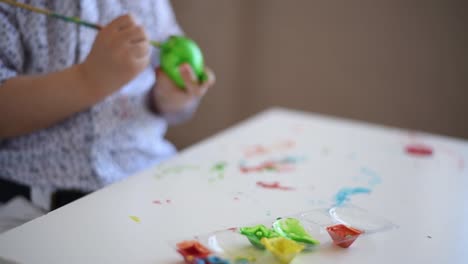 Die-Hände-eines-litlle-Kindes-mit-einem-Pinsel-malen-ein-Osterei-in-grüner-Farbe-auf-den-Tisch.