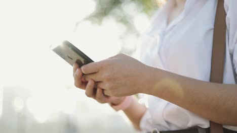 Close-up-hands-Asian-businesswoman-in-a-white-shirt-is-using-a-smartphone-texting-sharing-messages-on-social-media-while-outside-the-morning-at-a-public-park.