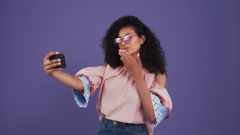 Señora-afroamericana-vestida-de-mujer-casual.-Disfrutaba-de-una-videollamada-en-su-smartphone,-sonriendo,-enviando-besos-aéreos.-Posando-sobre-fondo-púrpura.-De-cerca