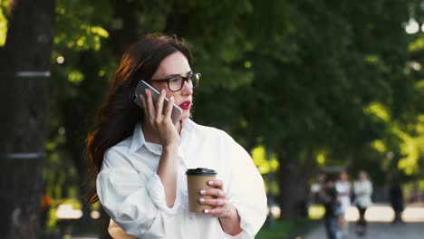 Mujer-con-gafas,-camisa-blanca.-Hablar-en-el-teléfono-inteligente-y-sostener-una-taza-de-café,-de-pie-en-el-parque-con-árboles-verdes.-Concepto-de-negocio.-Cámara-lenta