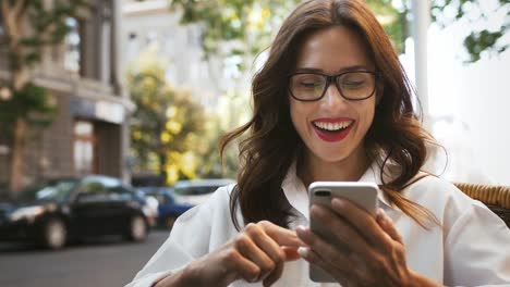 Business-girl-in-glasses,-white-shirt.-Looking-surprised-and-smiling,-reading-news-using-smartphone.-Sitting-in-outdoor-cafe.-Close-up,-slow-motion