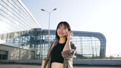 Good-looking-jocund-25-aged-asian-brunette-talking-on-mobile-near-airport-building-in-day-time