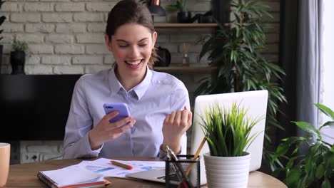 Divertido-y-eufórico-joven-mujer-enviando-mensajes-de-texto-en-el-teléfono-inteligente-en-la-oficina-en-casa.-Chica-emocionada-recibir-leer-buenas-noticias-en-el-teléfono-celular.