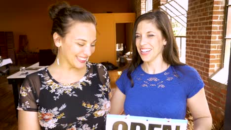 Portrait-of-two-beautiful-female-business-partners-hold-an-open-sign-and-smile