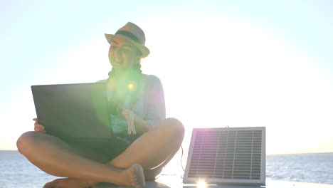 trip,-young-woman-sitting-on-car-roof-top-with-notebook-and-solar-panel-on-background-ocean-coast-in-backlit