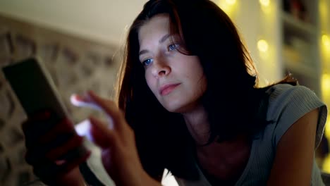 Close-up-of-young-concentrated-woman-having-internet-addiction-and-insomnia-using-smartphone-lying-in-bed-at-home-at-night