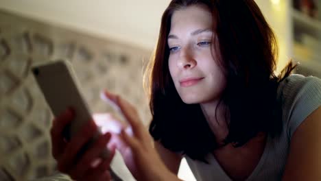 Close-up-of-young-smiling-woman-using-smartphone-lying-in-bed-at-home-at-night