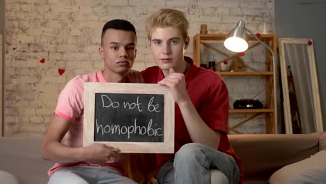 A-sad-international-gay-couple-is-sitting-on-the-couch-and-holding-a-sign.-Do-not-be-homophobic.-Look-at-the-camera.-Home-comfort-on-the-background.-60-fps