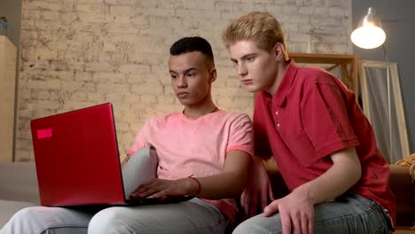 Two-multinational-homosexual-friends-sit-on-the-couch,-use-a-laptop,-watch-an-interesting-video-on-the-Internet,-surprised.-Home-cosiness,-family,-internet-concept.-60-fps